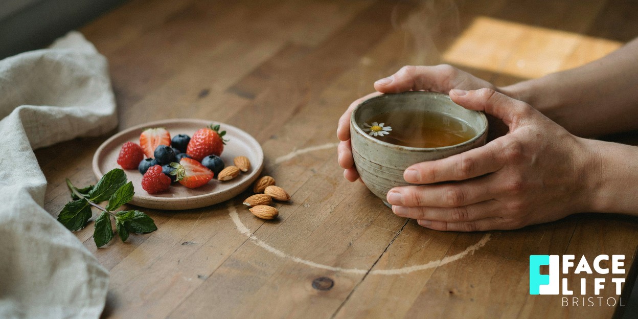A person’s hands cradling a warm herbal tea beside a small plate of berries and nuts in gentle morning light, suggesting a calmer, skin-friendly routine