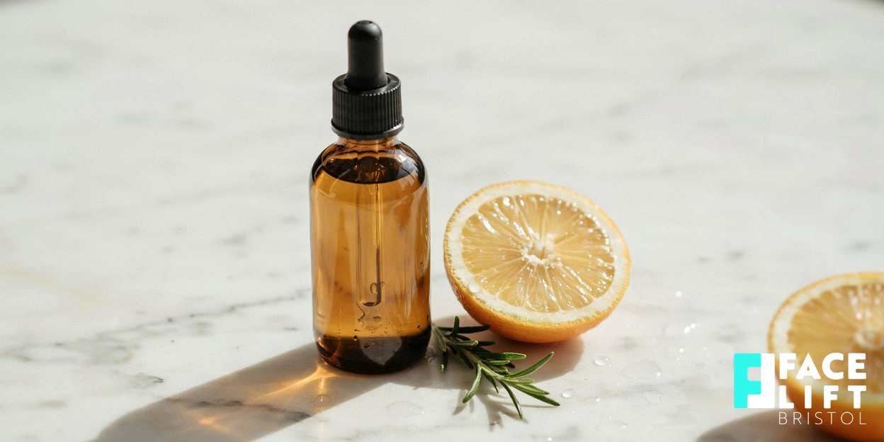 Editorial still-life of an amber glass dropper bottle beside a halved citrus fruit and a sprig of green botanical on a pale marble surface in soft daylight, evoking gentle acid-based skincare