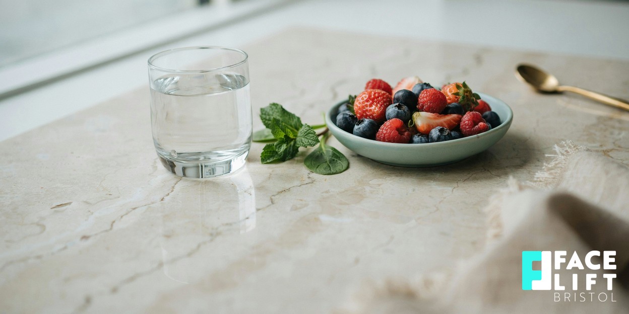 Calm wellness still-life of a glass of water, fresh berries and leafy greens arranged on a pale stone surface in soft daylight, evoking a skin-friendly lifestyle