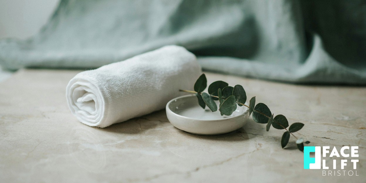 Calm modern clinic still-life with a rolled soft towel, a small ceramic dish and a sprig of greenery on a pale surface in warm daylight, evoking a hygienic, unhurried treatment room