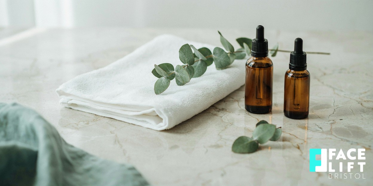 Calm modern clinic detail with a folded soft towel, a sprig of greenery and unbranded amber bottles on a stone surface in warm natural light