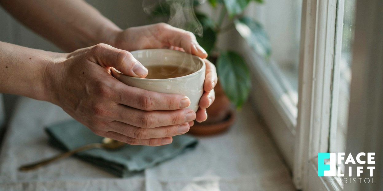 A person’s hands cradling a warm cup of herbal tea beside a sunlit window with a small green plant, evoking calm, unhurried self-care and healthy routine