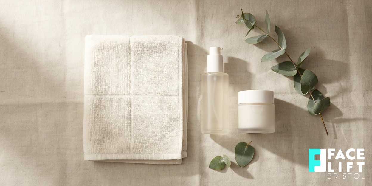 Overhead still-life of a calm desk scene with a folded soft towel, a sprig of eucalyptus and two unbranded skincare bottles arranged neatly on a warm neutral surface in soft daylight
