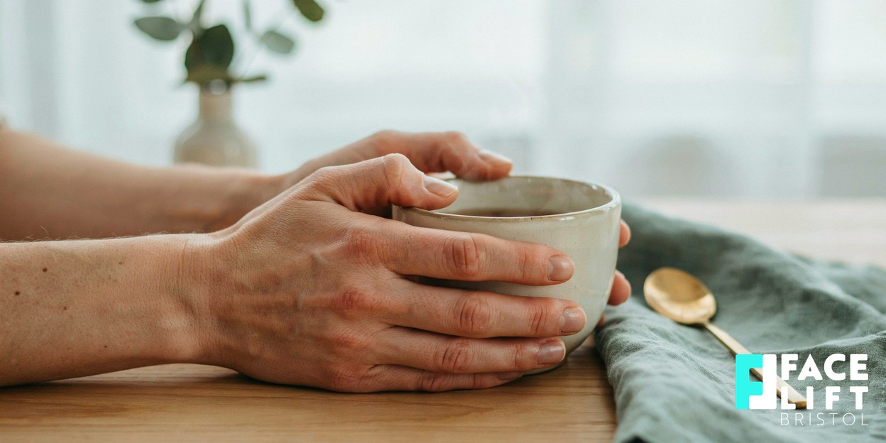 Close-up of a person’s hands resting calmly around a warm cup by a bright window, shallow depth of field, conveying an unhurried moment of considered self-care