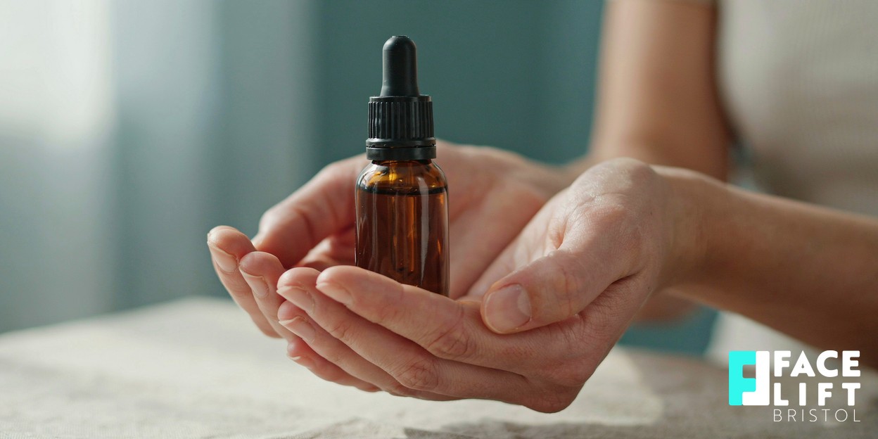 Close-up of a person’s hands gently holding a small amber glass skincare dropper bottle in warm natural daylight, conveying calm aftercare and self-care