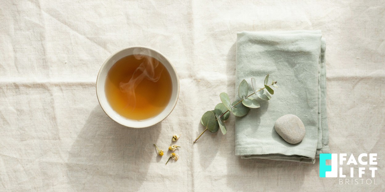 Overhead still life of a warm herbal tea, a folded soft towel and a sprig of greenery on a pale linen surface in gentle daylight, suggesting calm and stress relief