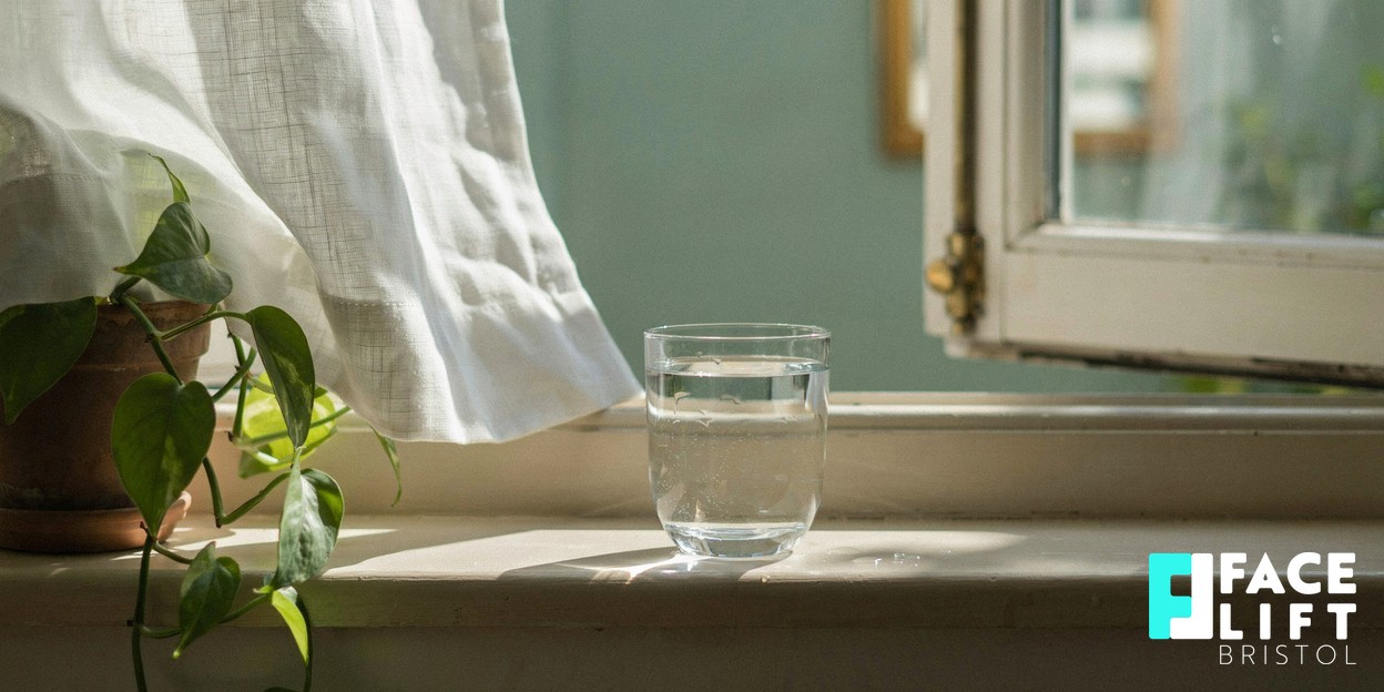 Photorealistic still life of a half-open window with a sheer curtain drifting in soft daylight and a glass of water on the sill, evoking fresh air, calm and a clean-living reset