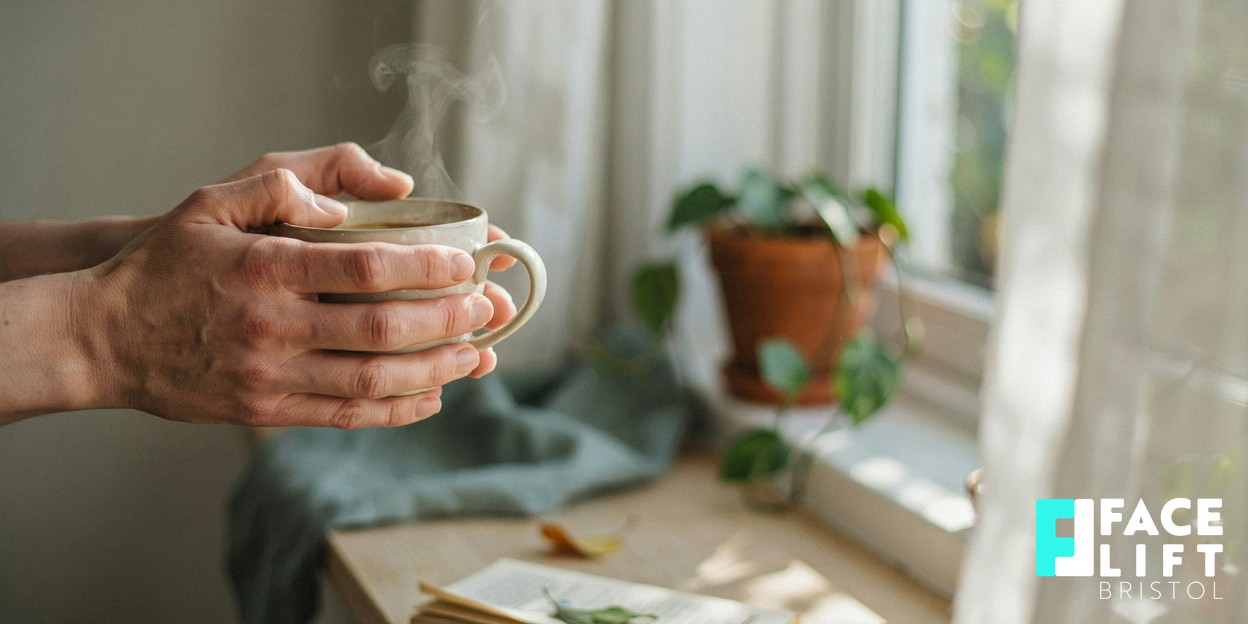 Person’s hands cradling a warm cup beside a window in soft natural daylight, a calm recovery moment conveying gentle downtime and self-care